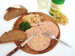 Holzteller mit Besteck, Saumagen, Brot, daneben eine Flasche Riesling und ein gefülltes Weinglas