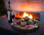 Holzbrett mit gegrillten Fleischspießen, einer Flasche und einem Glas Rotwein vor brennendem Kamin