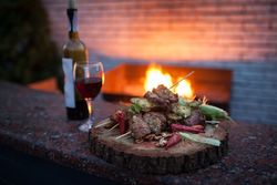 Holzbrett mit gegrillten Fleischspießen, einer Flasche und einem Glas Rotwein vor brennendem Kamin