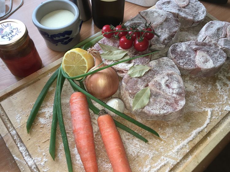 Holzbrett mit mehligen Kalbsbeinscheiben, zwei Karotten, einer Zwiebel, Tomaten, Knoblauch und Zitrone